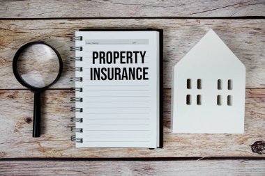 Kağıt defter üzerindeki mülkiyet sigortası metni beyaz ev modeli ve ahşap zemin üzerinde büyüteç