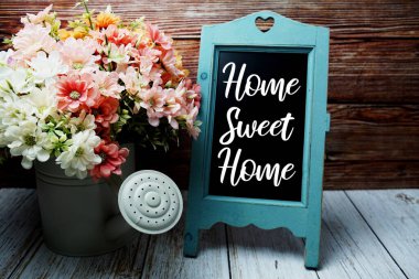 Home Sweet Home metni sehpada tahta zemin üzerinde metal vazo dekorasyonunda çiçek buketi bulunan karatahta
