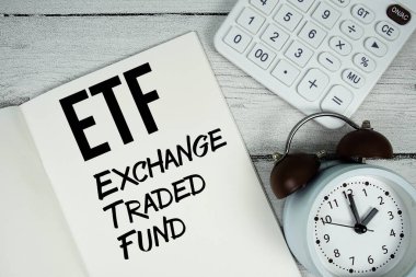 Kağıt defter sayfasında ETF Takas Fonu metni. Alarm saati ve tahta zemin üzerinde hesap makinesi var.