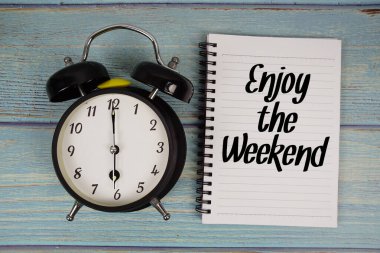Not defterine yazılan ve ahşap arka planda çalar saat olan Hafta sonu metninin tadını çıkar