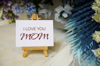 Seni seviyorum, Anneler Günün kutlu olsun. Kağıt karta tahta sehpa ve ahşap arka planda çiçek süslemesi yazılı.