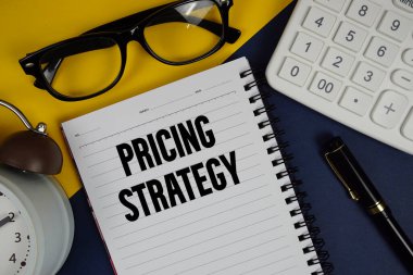 Not defterine yazılan fiyat stratejisi metni, hesap makinesi ve lacivert zemin üzerindeki alarm saati