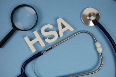 Sağlık Güvenliği alfabesinin kısaltılmışı. Mavi arka planda büyüteç ve steteskoplu harfler.