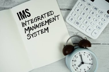 Kağıt defter üzerinde IMS Entegre Yönetim Sistemi metni. Alarm saati ve tahta zemin üzerinde hesap makinesi var.