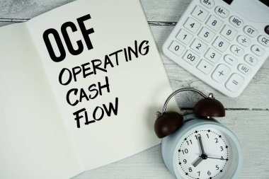 OCF, kağıt not defteri sayfasında nakit akışı metni işletiyor. Alarm saati ve ahşap zemin üzerinde hesap makinesi var.