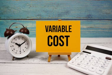 Kağıt kartta, hesap makinesi ve ahşap zemin üzerinde alarm saati ile yazılmış değişken maliyet metni