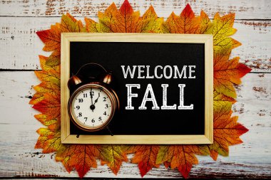 Ahşap zemin üzerine akçaağaç yaprağı süslenmiş alarm saati ile sonbahar metnini hoş geldiniz