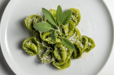 Beyaz arkaplanda adaçayı ve parmesanlı yeşil ravioli tabağının üst görüntüsü.