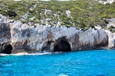 Mavi gökyüzü, dağları, kayaları, denizi ve turkuaz suyu olan kayalık sahil Yunanistan 'daki Zakynthos Adası' nın arka planında.
