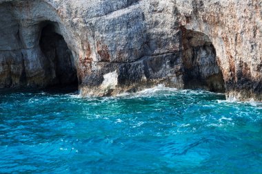 Mavi gökyüzü, dağları, kayaları, denizi ve turkuaz suyu olan kayalık sahil Yunanistan 'daki Zakynthos Adası' nın arka planında.