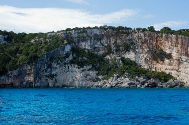 Mavi gökyüzü, dağları, kayaları, denizi ve turkuaz suyu olan kayalık sahil Yunanistan 'daki Zakynthos Adası' nın arka planında.