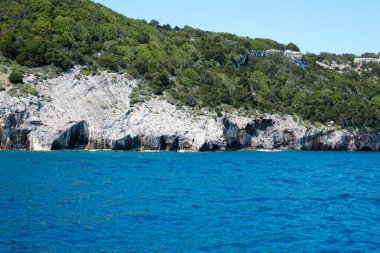Mavi gökyüzü, dağları, kayaları, denizi ve turkuaz suyu olan kayalık sahil Yunanistan 'daki Zakynthos Adası' nın arka planında.