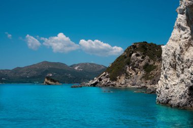 Mavi gökyüzü, dağları, kayaları, denizi ve turkuaz suyu olan kayalık sahil Yunanistan 'daki Zakynthos Adası' nın arka planında.