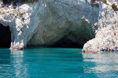 Mavi gökyüzü, dağları, kayaları, denizi ve turkuaz suyu olan kayalık sahil Yunanistan 'daki Zakynthos Adası' nın arka planında.