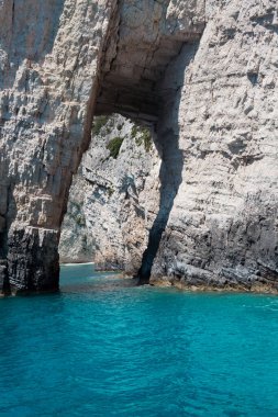 Mavi gökyüzü, dağları, kayaları, denizi ve turkuaz suyu olan kayalık sahil Yunanistan 'daki Zakynthos Adası' nın arka planında.