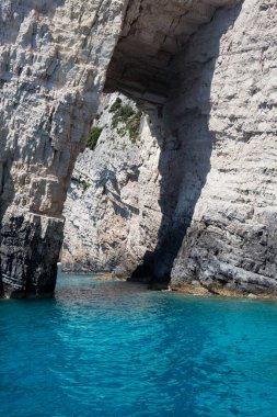 Mavi gökyüzü, dağları, kayaları, denizi ve turkuaz suyu olan kayalık sahil Yunanistan 'daki Zakynthos Adası' nın arka planında.