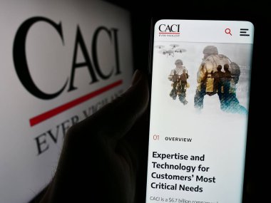 Stuttgart, Almanya - 04-28-2024: ABD 'li bilgi teknolojileri şirketi CACI International Inc.' in logolu web sayfası ile cep telefonu tutan kişi. Telefon ekranının merkezine odaklan.