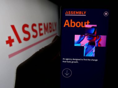 Stuttgart, Almanya - 05-16-2024: ABD 'li şirketin PMX Ajansı LLC (Assembly Global) tanıtım sayfası ile logolu cep telefonu tutan kişi. Telefon ekranının merkezine odaklan.