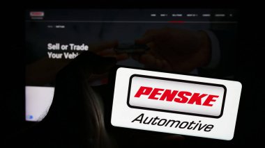 Stuttgart, Germany - 05-23-2024: Person holding mobile phone with logo of American dealership company Penske Automotive Group Inc. in front of web page. Focus on phone display. clipart