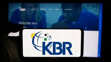 Stuttgart, Germany - 05-27-2024: Person holding cellphone with logo of US engineering and construction company KBR Inc. in front of business webpage. Focus on phone display. clipart