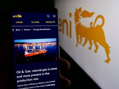 Stuttgart, Germany - 06-01-2024: Person holding cellphone with web page of Italian energy company Eni S.p.A. in front of business logo. Focus on center of phone display. clipart