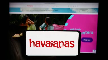 Stuttgart, Germany - 06-06-2024: Person holding cellphone with logo of Brazilian flip-flop manufacturing company Havaianas in front of business webpage. Focus on phone display. clipart