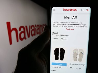 Stuttgart, Germany - 06-06-2024: Person holding cellphone with website of Brazilian flip-flop manufacturing company Havaianas in front of logo. Focus on center of phone display. clipart