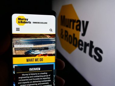 Stuttgart, Germany - 06-11-2024: Person holding cellphone with webpage of South African company Murray and Roberts Holdings Ltd. with logo. Focus on center of phone display. clipart