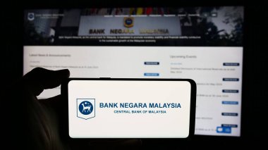 Stuttgart, Germany - 06-24-2024: Person holding cellphone with logo of Malaysian institution Bank Negara Malaysia (BNM) in front of webpage. Focus on phone display. clipart