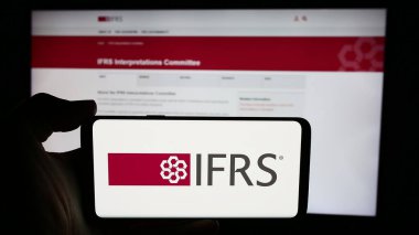 Stuttgart, Germany - 06-26-2024: Person holding cellphone with logo of International Financial Reporting Standards Foundation (IFRS) in front of webpage. Focus on phone display. clipart