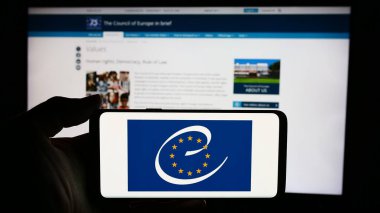 Stuttgart, Germany - 06-26-2024: Person holding cellphone with logo of human rights organisation Council of Europe (CoE) in front of webpage. Focus on phone display. clipart