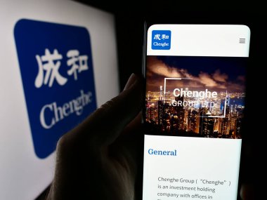 Stuttgart, Almanya - 06-26-2024: Yatırım şirketi Chenghe Group Ltd. 'nin web sitesi ile iş logosu önünde cep telefonu bulunduran kişi. Telefon ekranının merkezine odaklan.