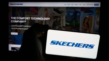 Stuttgart, Almanya - 06-27-2024: Amerikan ayakkabı şirketi Skechers USA Inc. 'in logosu olan cep telefonu sahibi kişi iş sayfasının önünde. Telefon ekranına odaklan.