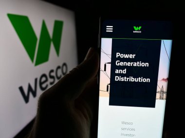 Stuttgart, Almanya - 07-05-2024: ABD 'li elektronik şirketi WESCO International Inc.' nin internet sayfası ile logo önünde cep telefonu tutan kişi. Telefon ekranının merkezine odaklan.