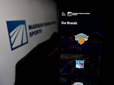 Stuttgart, Germany - 07-06-2024: Person holding cellphone with webpage of US company Madison Square Garden Sports Corp. in front of logo. Focus on center of phone display. clipart