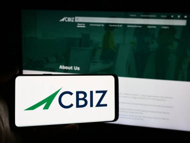 Stuttgart, Almanya - 07-06-2024: Amerikan finans danışmanlık şirketi CBIZ Inc. 'in logosuna sahip cep telefonu sahibi kişi iş sayfasının önünde. Telefon ekranına odaklan.