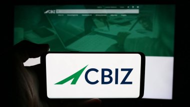 Stuttgart, Almanya - 07-06-2024: ABD 'li finans danışmanlık şirketi CBIZ Inc.' in logosuyla cep telefonu sahibi kişi iş sayfası önünde. Telefon ekranına odaklan.