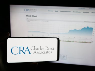 Stuttgart, Germany - 07-06-2024: Person holding mobile phone with logo of CRA International Inc. (Charles River Associates) in front of company web page. Focus on phone display. clipart