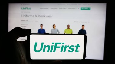 Stuttgart, Almanya - 07-06-2024: ABD 'li koruyucu giyim şirketi UniFirst Corporation' ın logosuyla cep telefonu sahibi kişi iş sayfası önünde. Telefon ekranına odaklan.