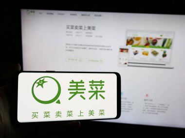 Stuttgart, Almanya - 07-10-2024: İnternet sitesi önünde Çinli tarım e-ticaret şirketi Meicai 'nin logosuna sahip akıllı telefon sahibi kişi. Telefon ekranına odaklan.