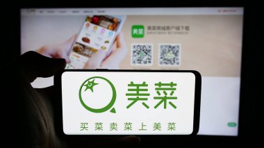 Stuttgart, Germany - 07-10-2024: Person holding cellphone with logo of Chinese agricultural e-commerce company Meicai in front of business webpage. Focus on phone display. clipart