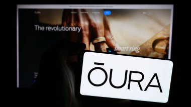 Stuttgart, Germany - 07-11-2024: Person holding mobile phone with logo of Finnish health technology company Oura Health Oy in front of business web page. Focus on phone display. clipart