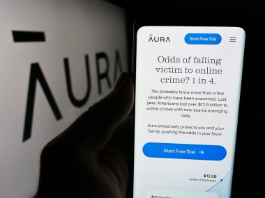Stuttgart, Almanya - 07-12-2024: ABD 'li siber güvenlik yazılım şirketi Aura Sub LLLC' nin web sitesi ile logo önünde cep telefonu tutan kişi. Telefon ekranının merkezine odaklan.