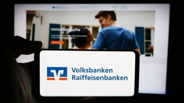 Stuttgart, Almanya - 07-15-2024: Alman bankacılık ağı Volksbanken Raiffeisenbanken 'in logosuna sahip cep telefonu sahibi kişi iş sayfası önünde. Telefon ekranına odaklan.