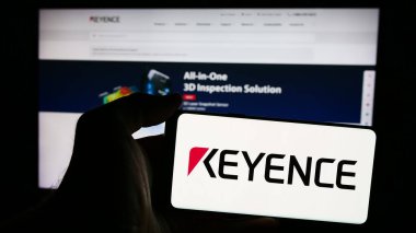 Stuttgart, Almanya - 07-17-2024: Japon elektronik şirketi Keyence Corporation 'ın logosunun bulunduğu cep telefonu sahibi kişi iş sayfasının önünde. Telefon ekranına odaklan.