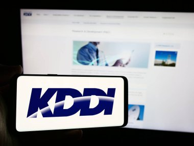 Stuttgart, Almanya - 07-19-2024: Japon telekomünikasyon şirketi KDDI Corporation 'ın logosunun bulunduğu cep telefonu sahibi kişi web sayfası önünde. Telefon ekranına odaklan.