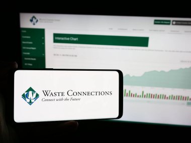 Stuttgart, Germany - 07-19-2024: Person holding mobile phone with logo of American recycling company Waste Connections Inc. in front of business web page. Focus on phone display. clipart