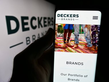 Stuttgart, Germany - 07-23-2024: Smartphone with website of US footwear company Deckers Outdoor Corporation in front of business logo. Focus on center of phone display. clipart