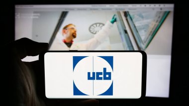 Stuttgart, Almanya - 07-25-2024: Belçikalı biyofarmasötik şirketi UCB S.A. 'nın logosuyla cep telefonu tutan kişi iş sayfasının önünde. Telefon ekranına odaklan.