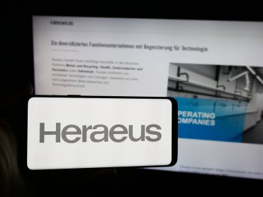 Stuttgart, Germany - 07-31-2024: Person holding smartphone with logo of German technology company Heraeus Holding GmbH in front of website. Focus on phone display. clipart